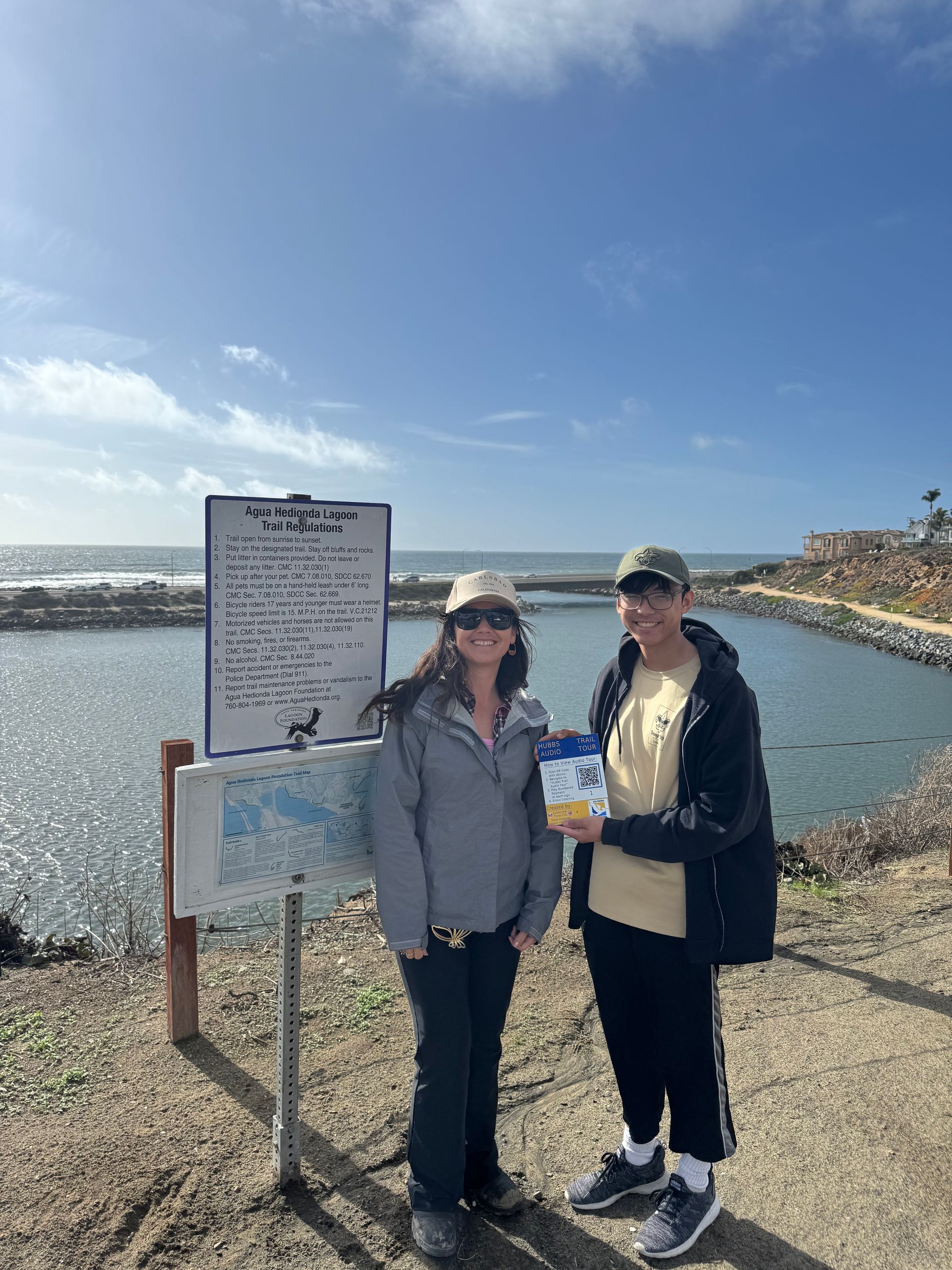 Two people stand by a sign and water. One holds a blue and white package. Sunny day, blue sky.