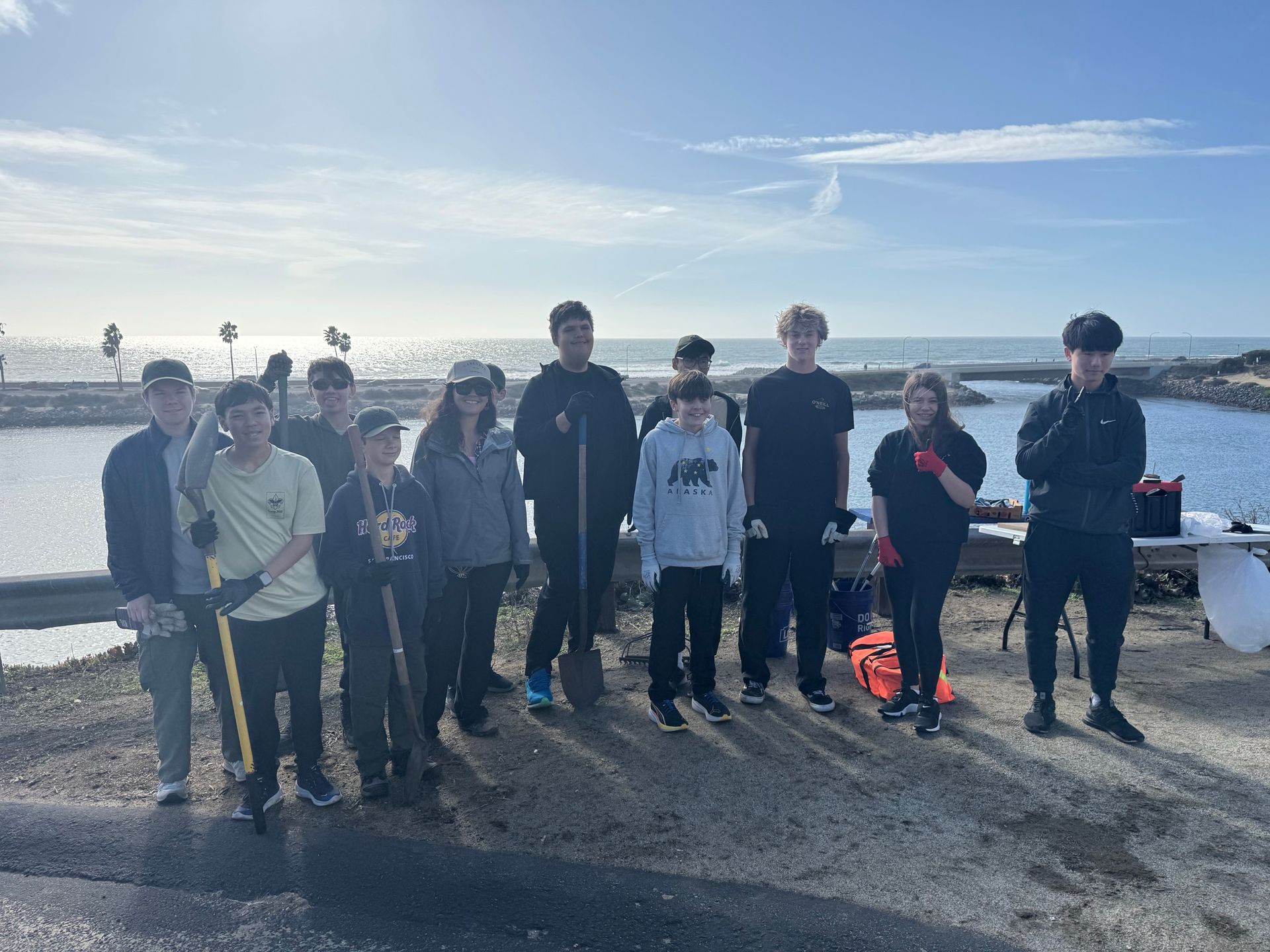 Group of people standing on a stone structure near ocean, holding tools. Blue sky, sunny.