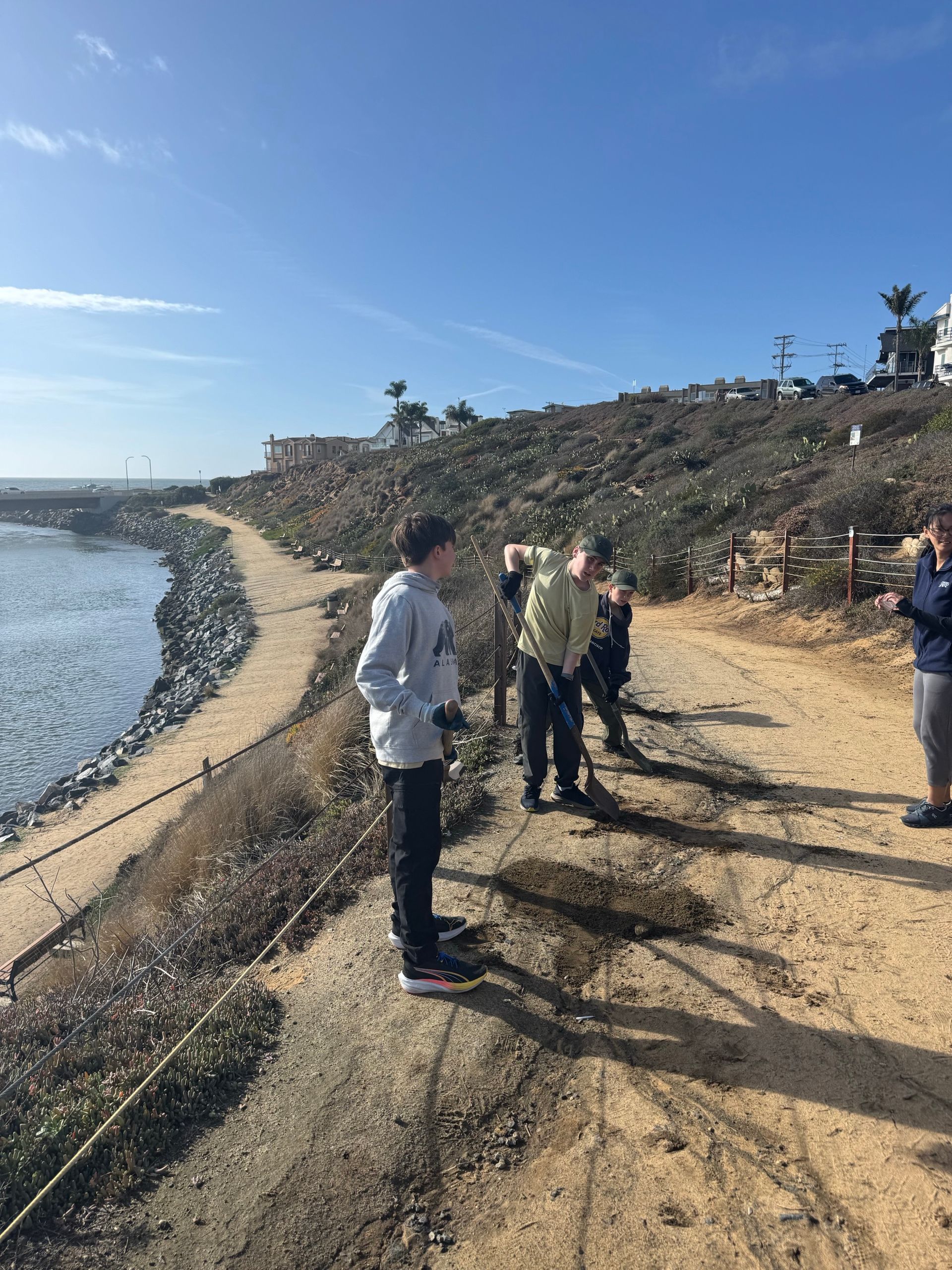 People working on a dirt path along a coast, blue water and sky.