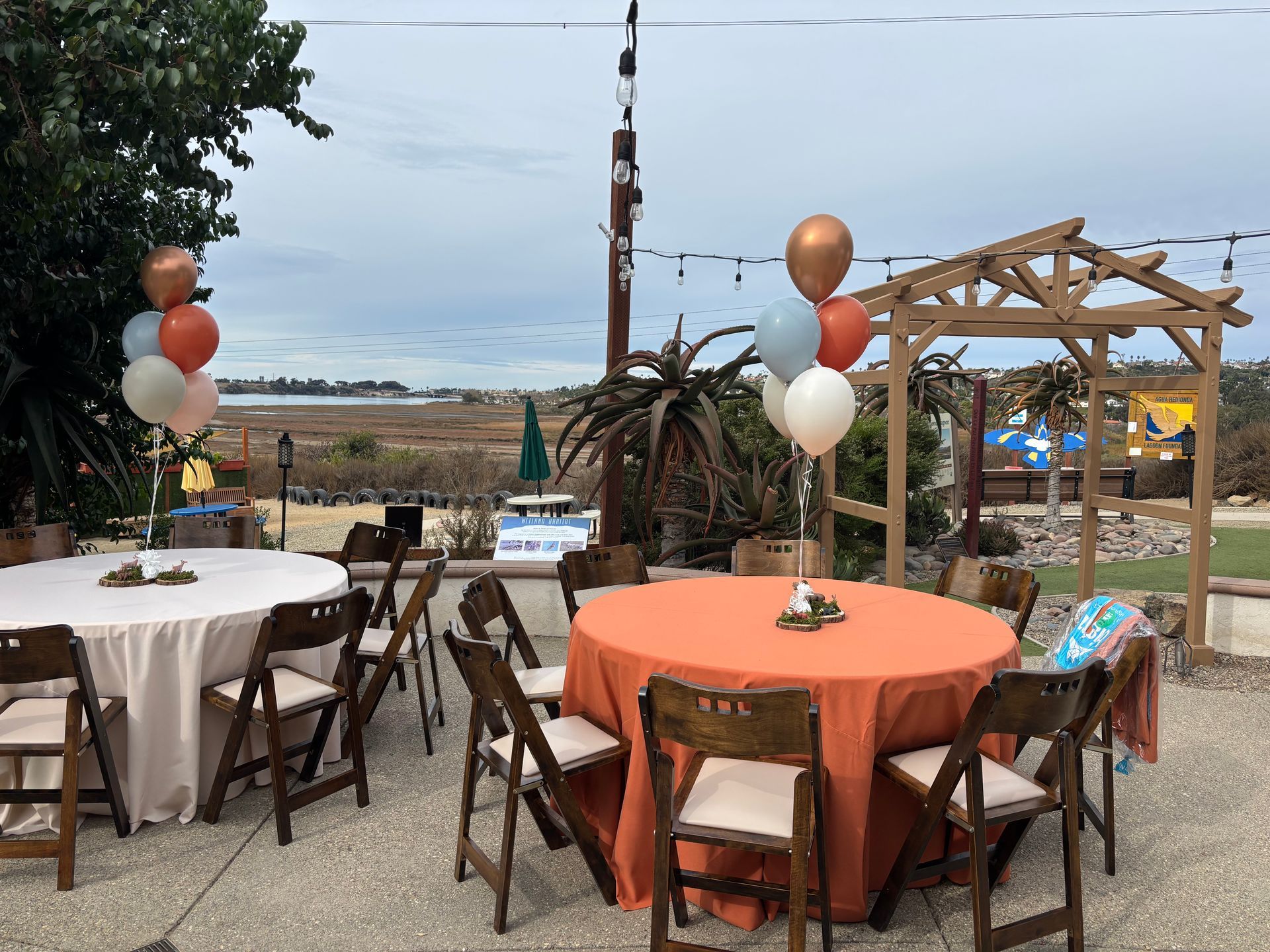 Outdoor event setup with round tables, chairs, balloons, and a wooden arbor. Overcast sky and marsh views.
