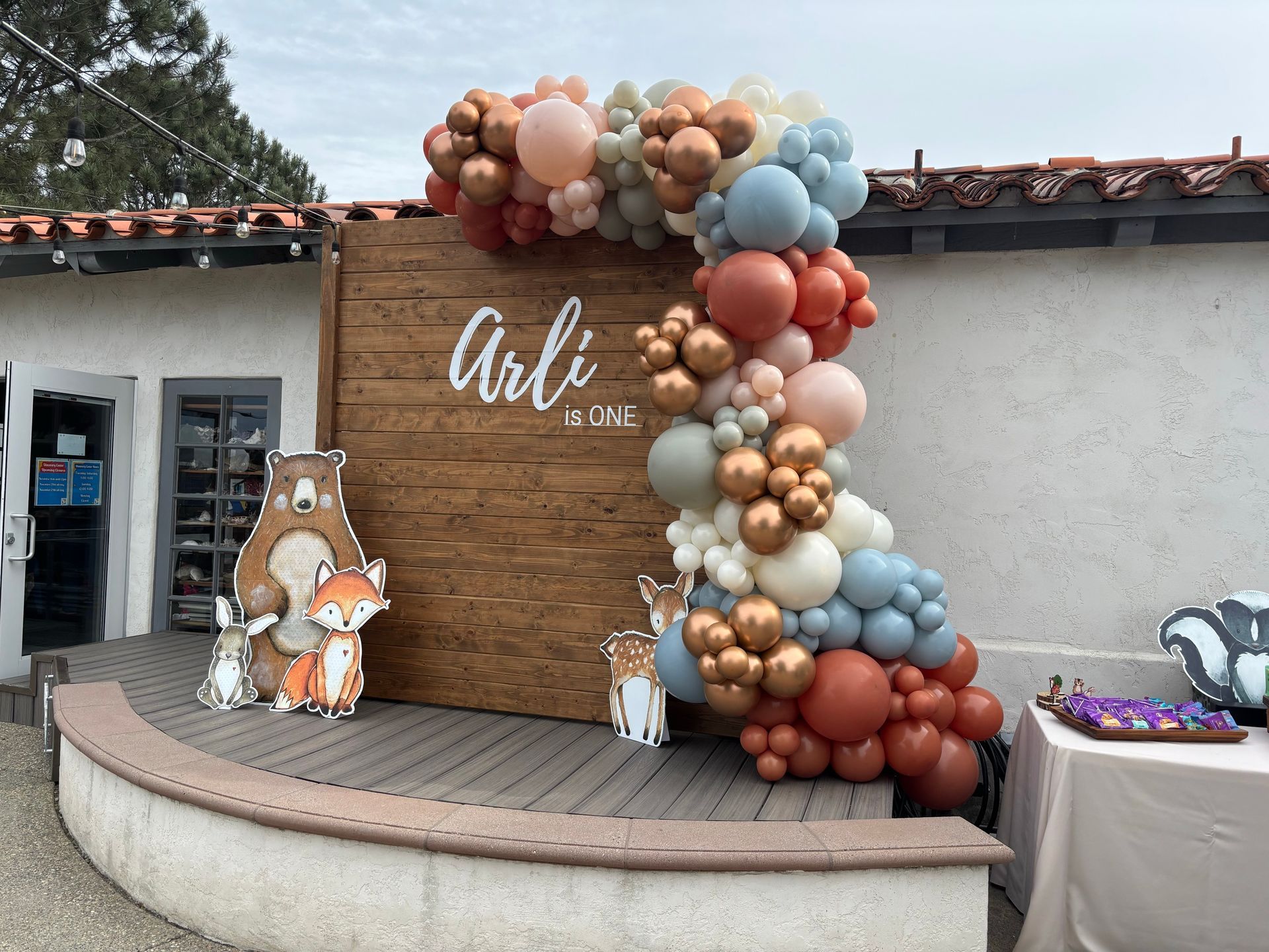 Balloon arch with wooden backdrop for an event. Woodland animal cutouts and a table with treats are visible.