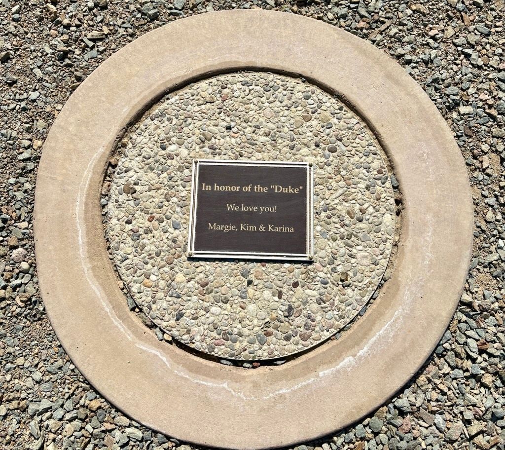 Bronze plaque within a concrete ring on gravel, reads: 