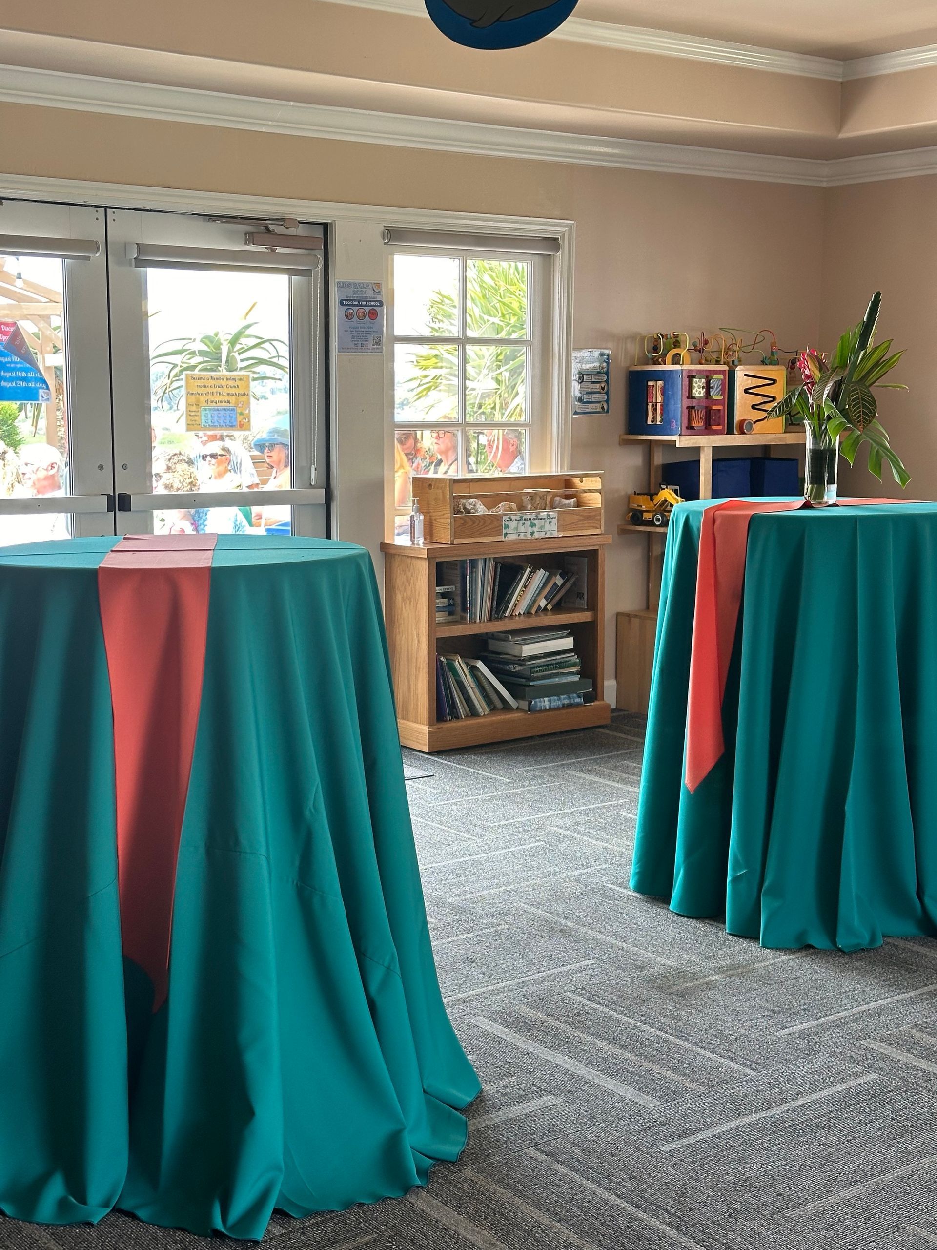 Two round tables with teal cloths and coral runners, inside a room with a window, shelves, and a door.