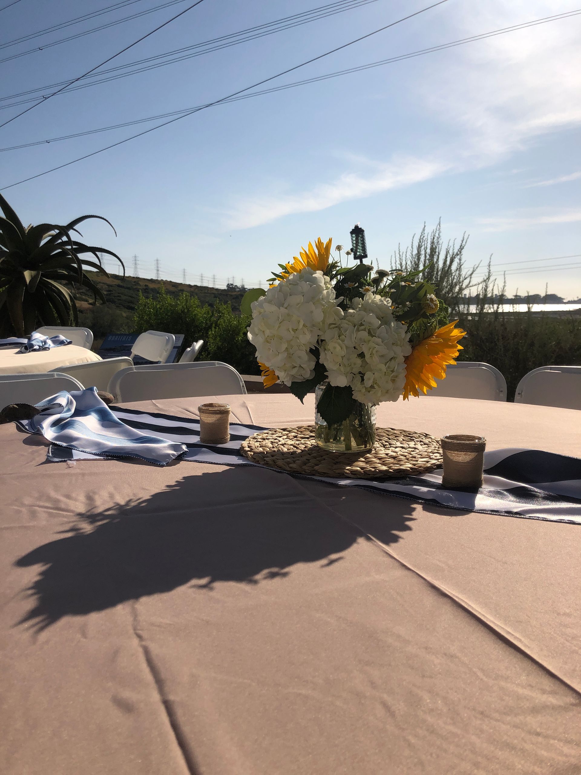 Table setting with floral arrangement, outdoor wedding. Sunflowers, hydrangeas, candles, blue runner, beige tablecloth.