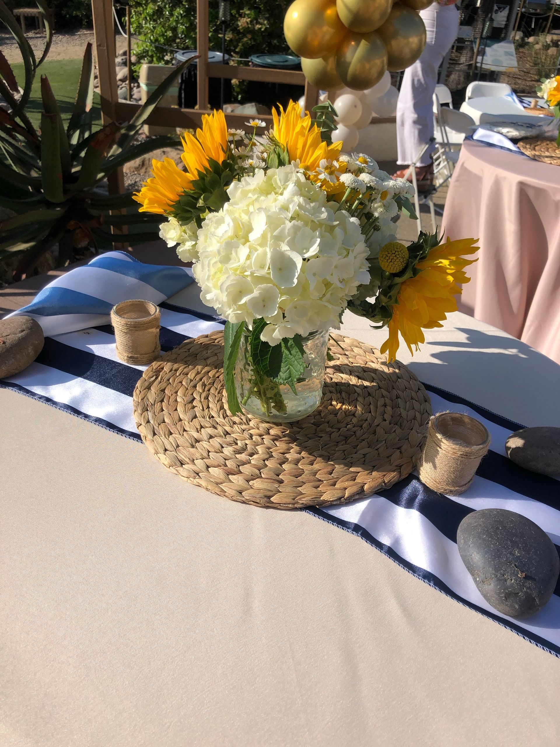 Centerpiece with sunflowers, white hydrangeas, on a table set with a striped runner and decorative rocks.