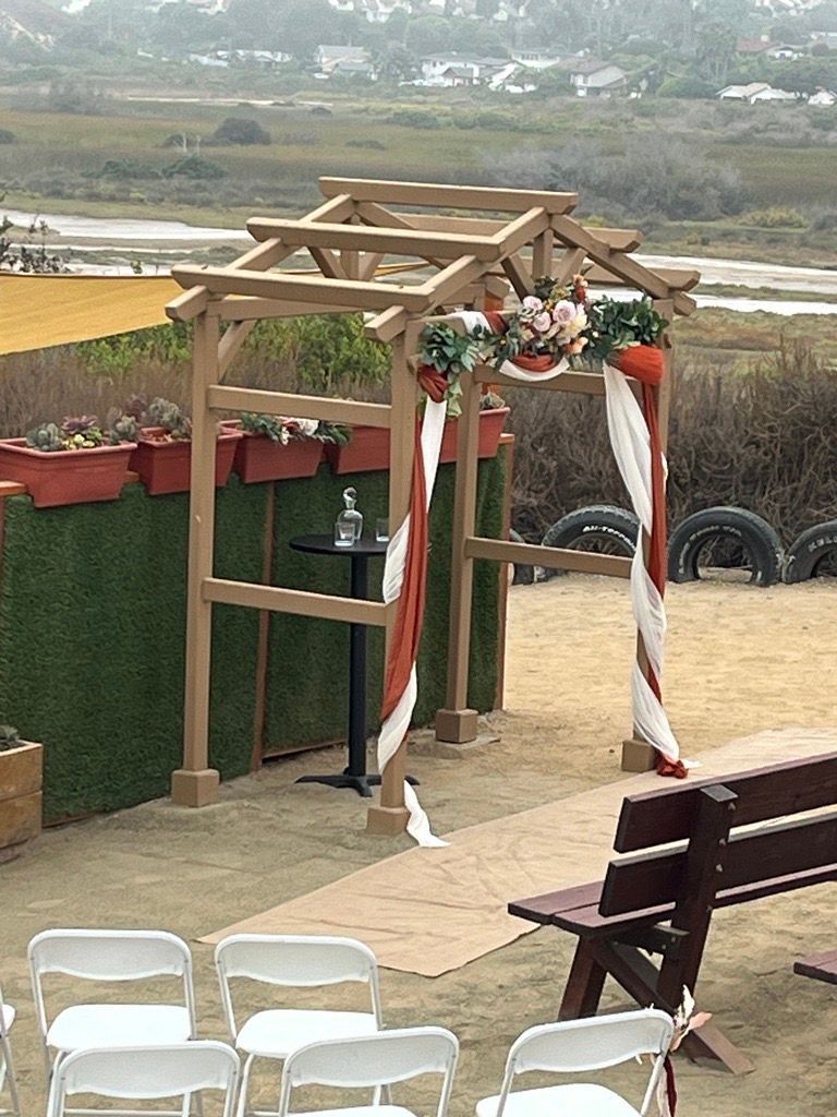 Wedding arch decorated with flowers and fabric, in front of greenery and a brown bench.