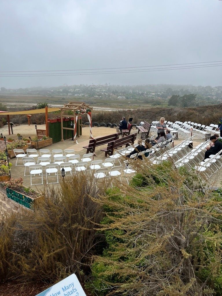 Outdoor wedding setup on a foggy day. Rows of white chairs face an altar, with guests and a landscape backdrop.