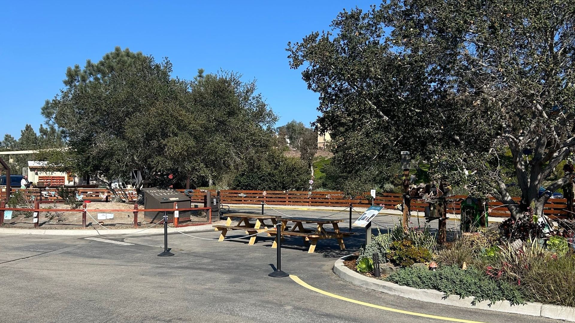 Entrance to a park with trees, picnic tables, and a wooden fence on a sunny day.