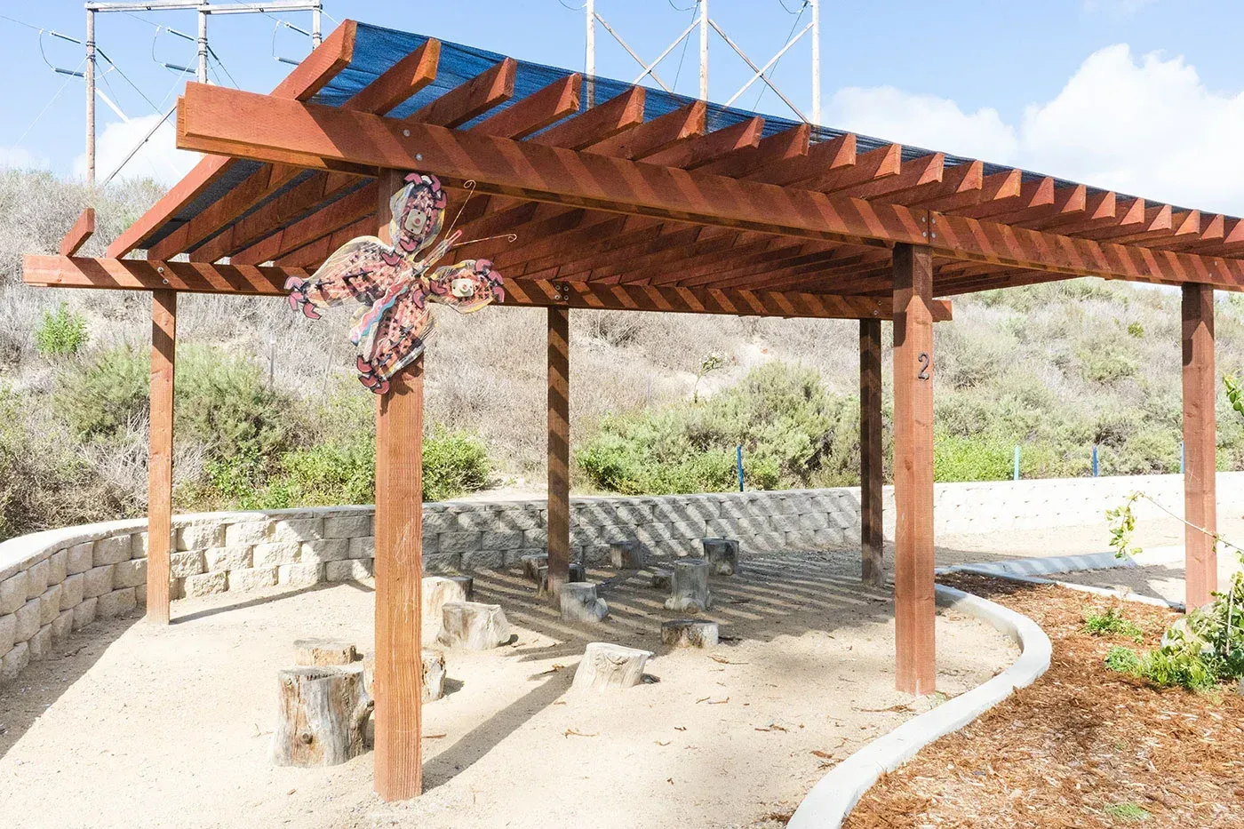 Wooden pergola with a dragonfly sculpture in a sunny outdoor setting with a gravel base.