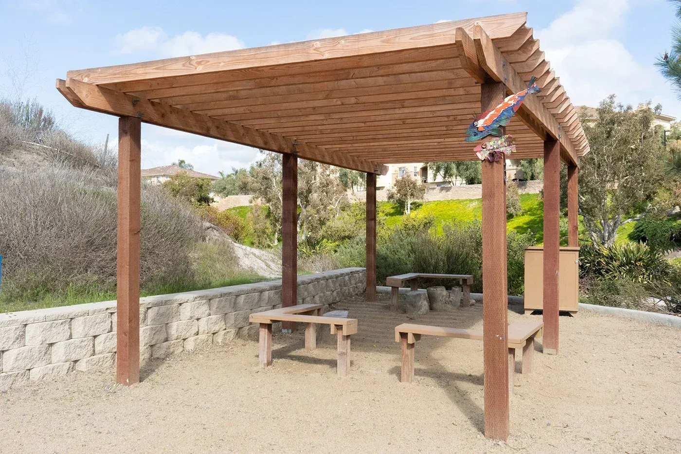 Wooden pergola over a gravel area with benches, a fire pit, and a trash can.