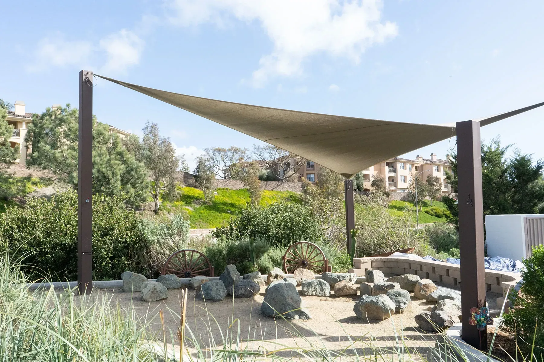 Shade sail over a rock garden, brown posts, greenery, and buildings in the background. Sunny day.