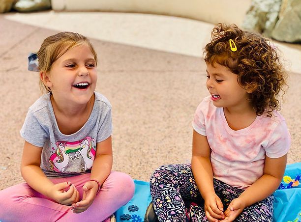 Two young girls laughing while sitting on a blanket, one wearing pink leggings, the other a tie-dye shirt.