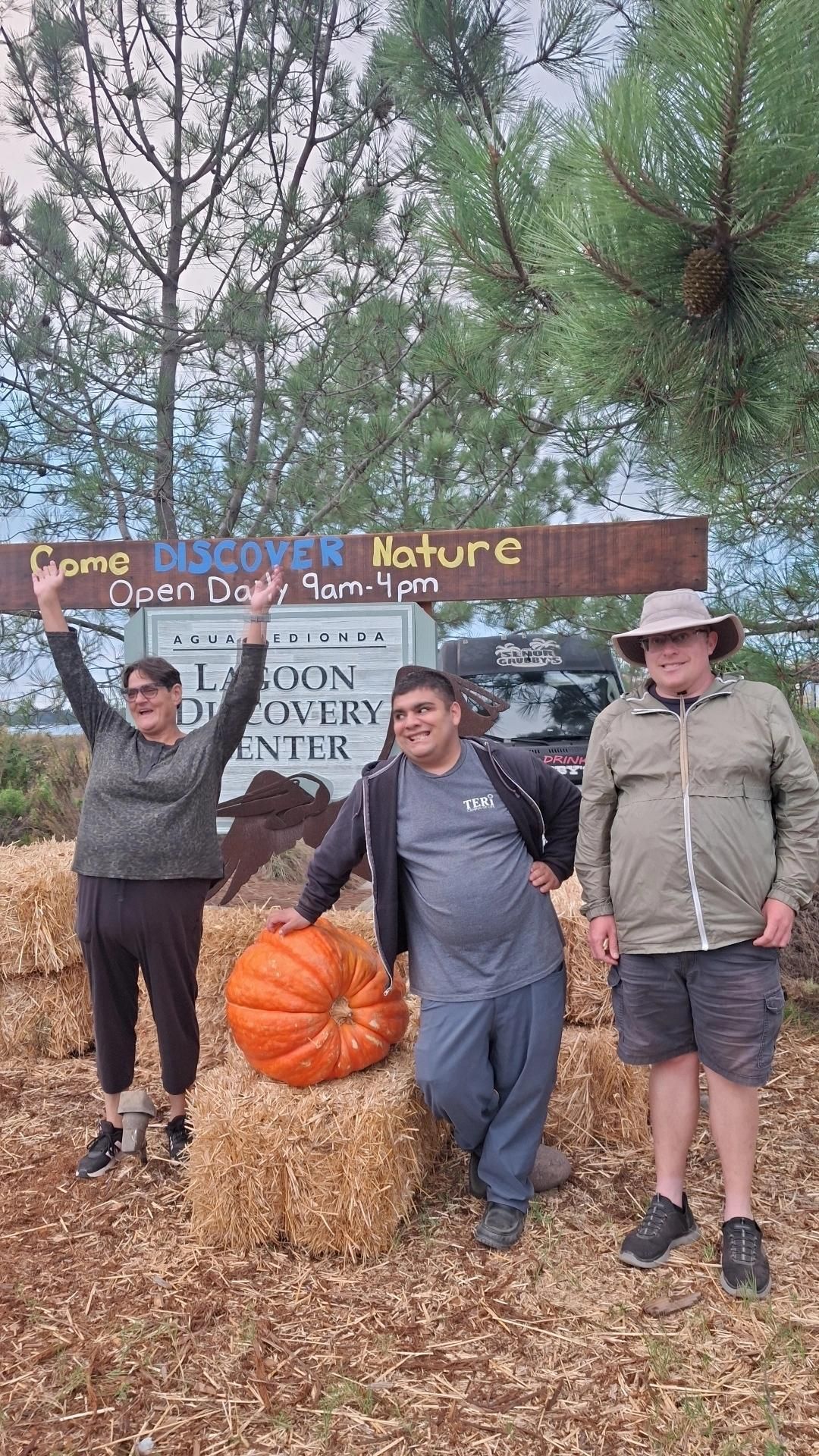 Three people stand near a pumpkin and a sign. The sign reads 