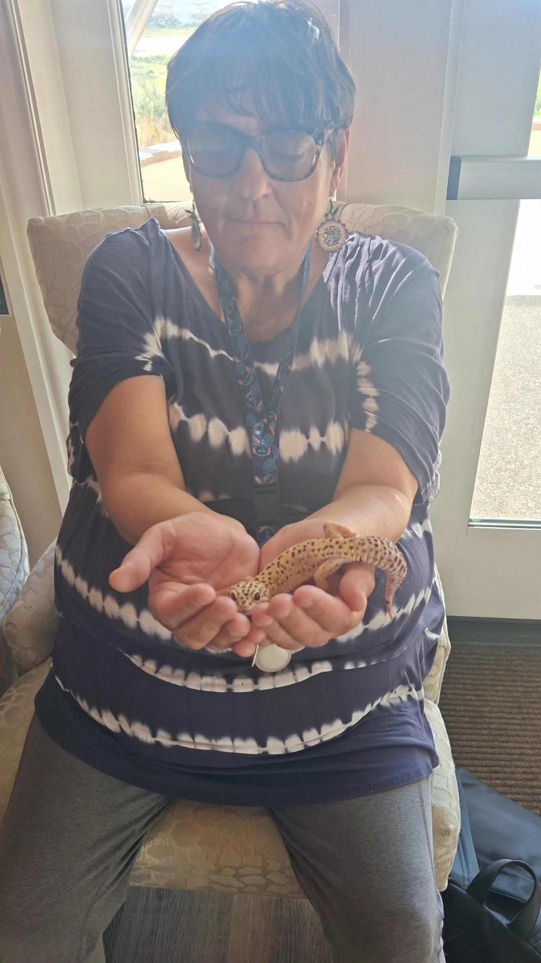 Woman holding a gecko in her hands; she is wearing a blue tie-dye shirt and glasses; indoors.