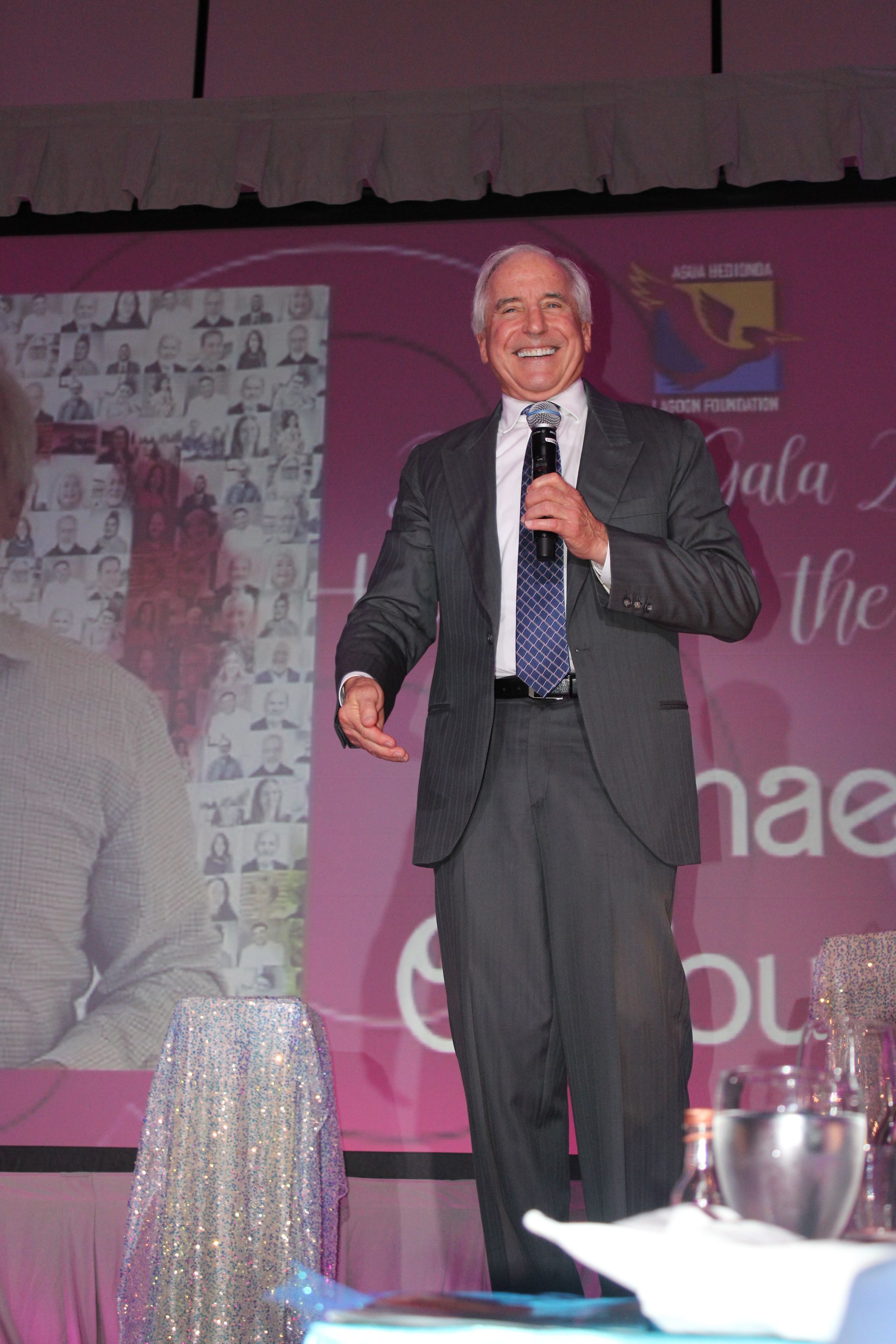 Man in a dark suit speaking into a microphone on a pink-lit stage with event banners behind him