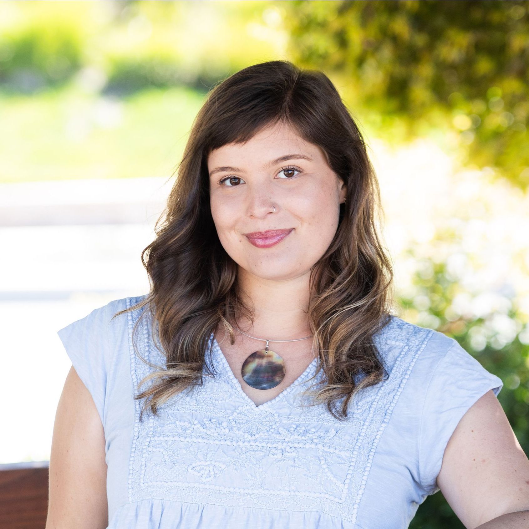 Woman with brown hair and a light blue shirt smiles outdoors.