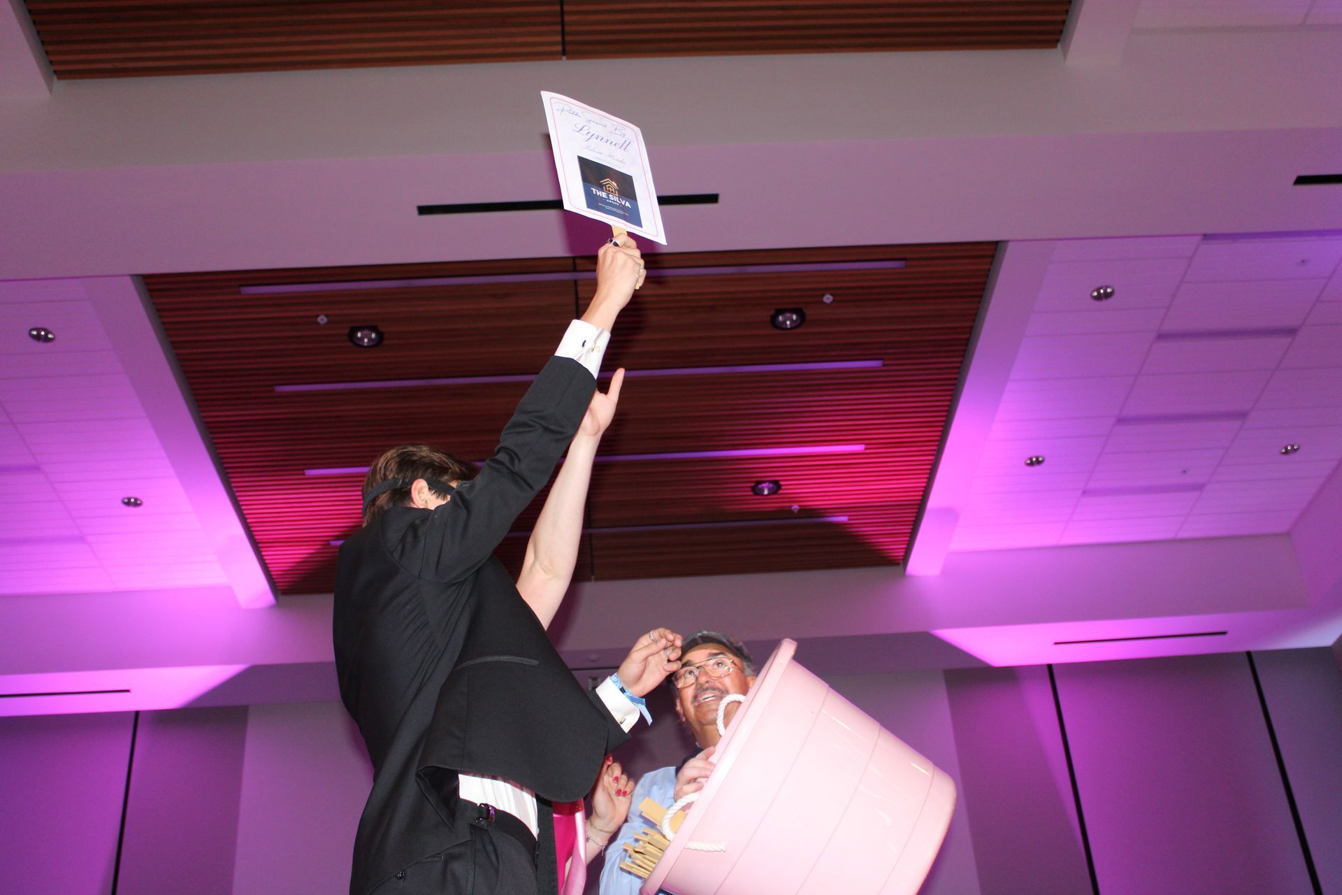 Two people reach up toward a sign at a purple-lit indoor event, one holding it overhead.