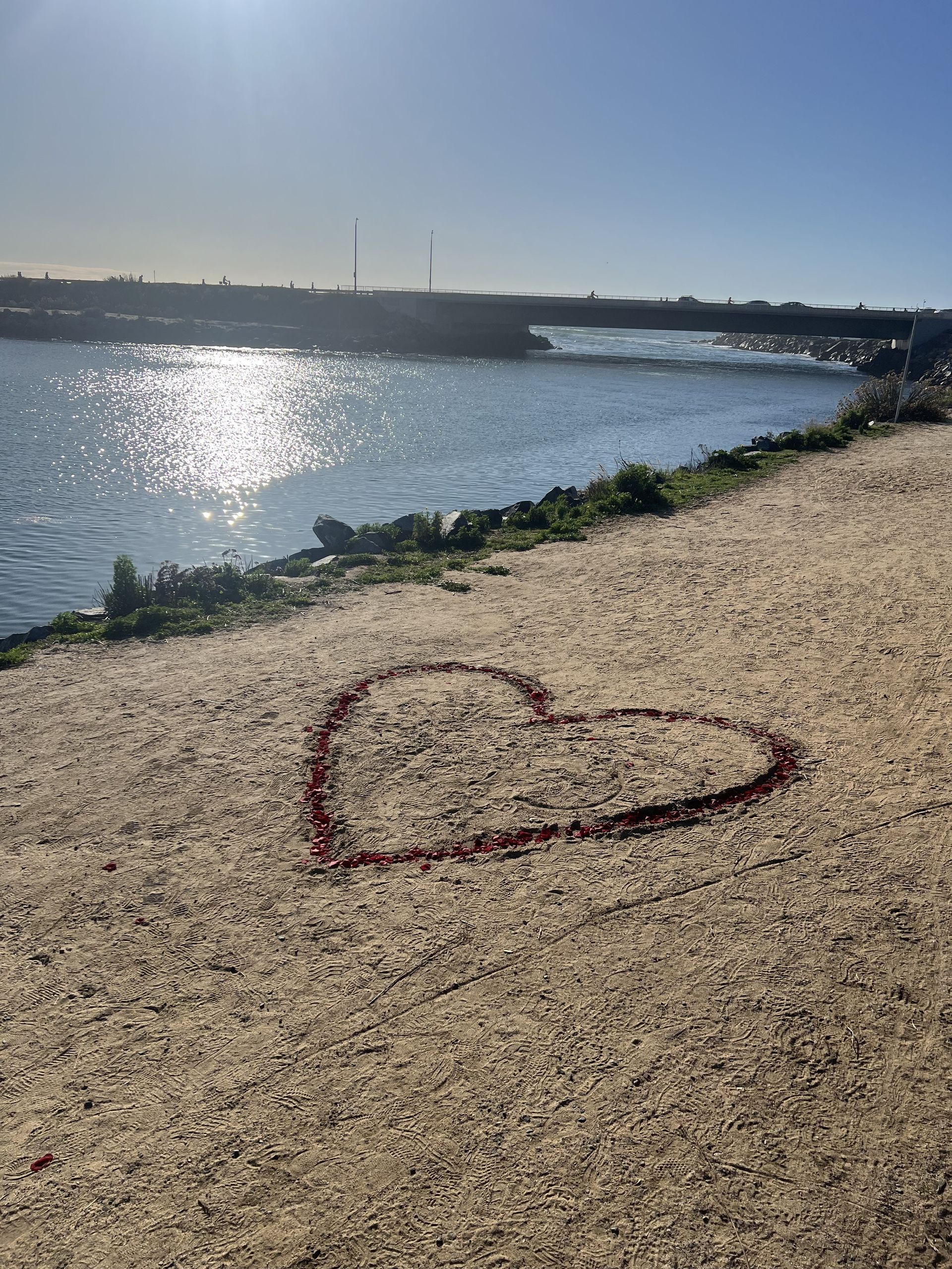 Heart drawn in red on a sandy path, overlooking water. Sunlight glistens.