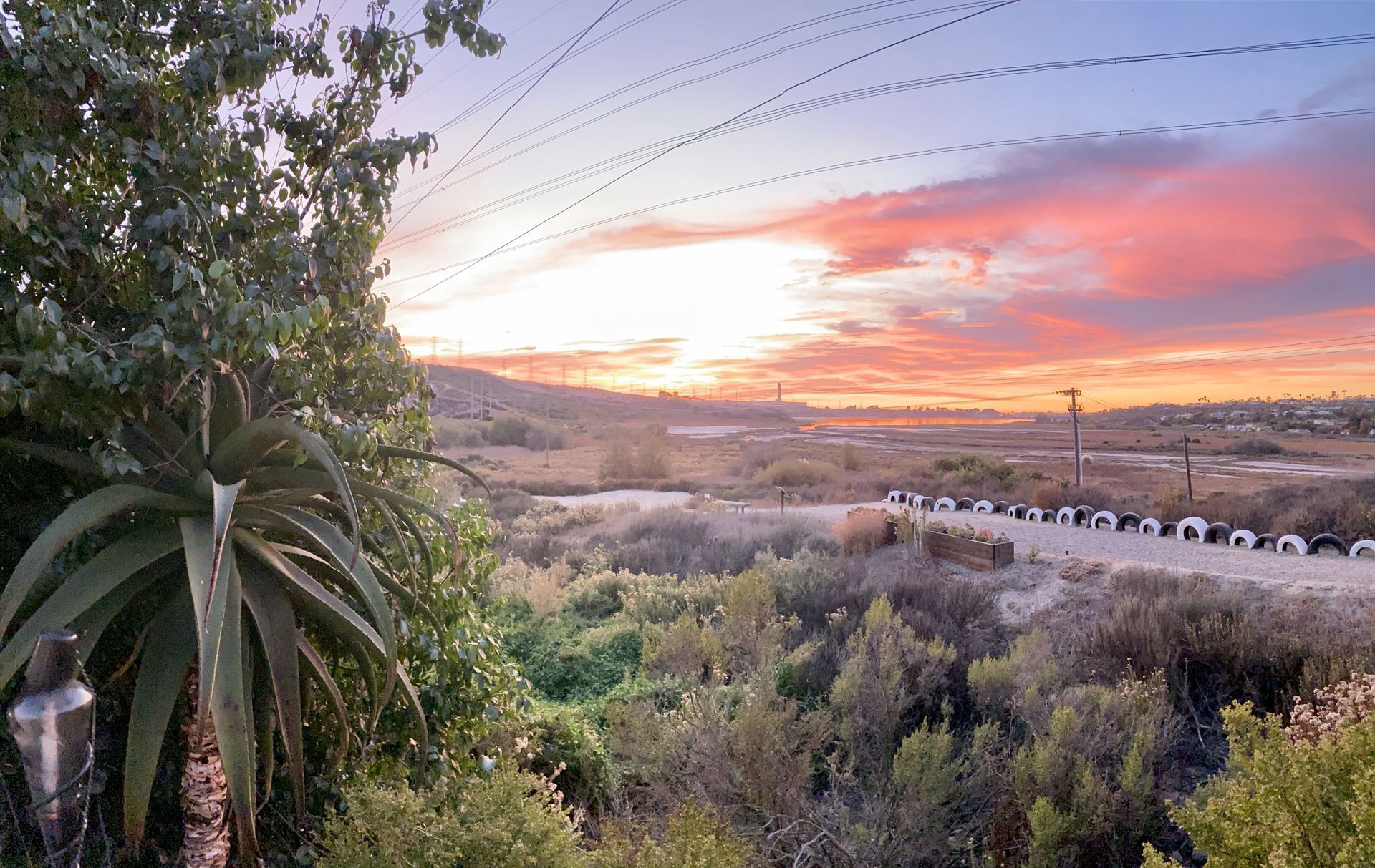 Sunset over a valley with green foliage, a road, and power lines. Sky is pink and orange.