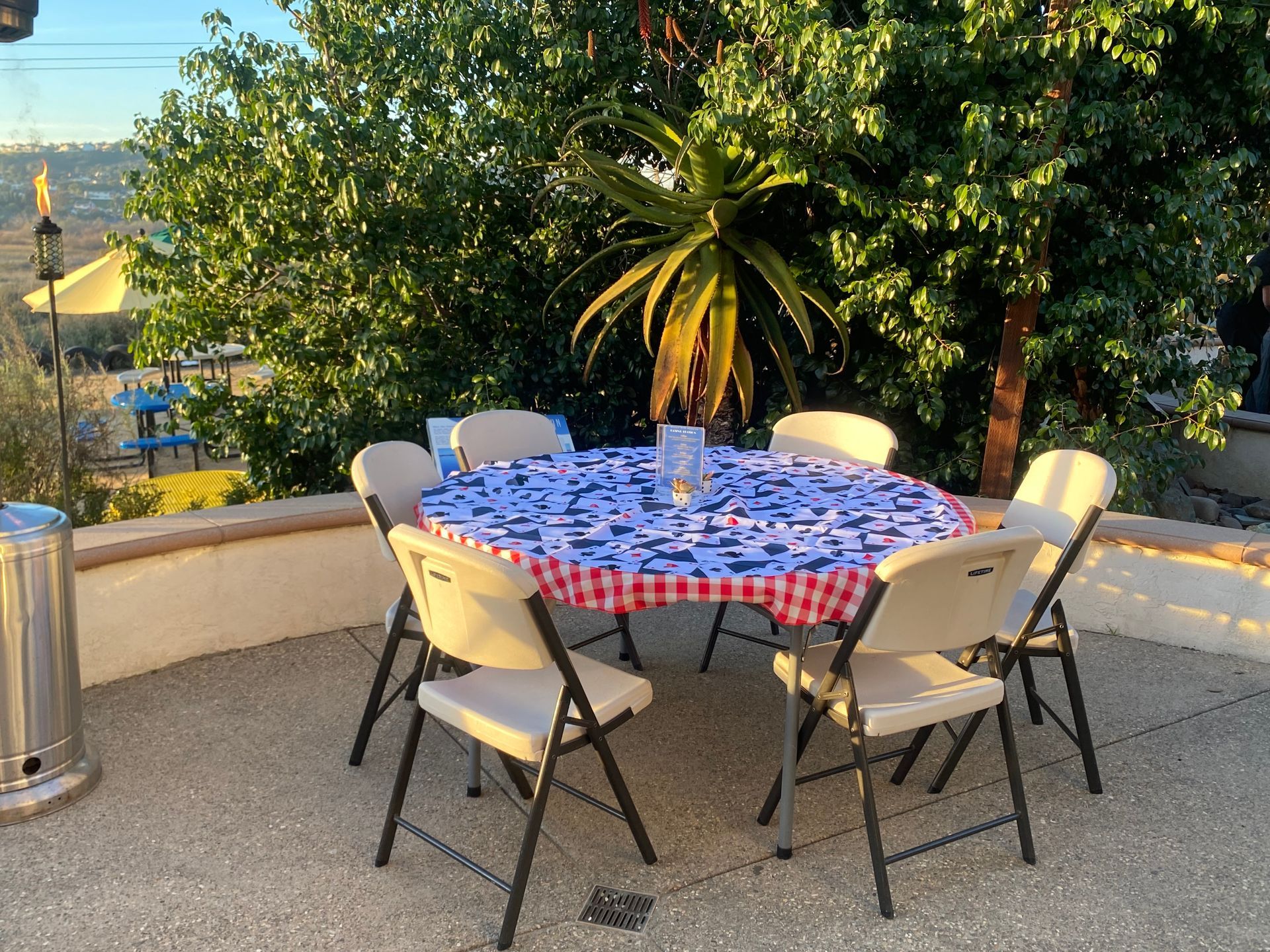 A round table with a blue and red patterned tablecloth and six folding chairs on an outdoor patio.