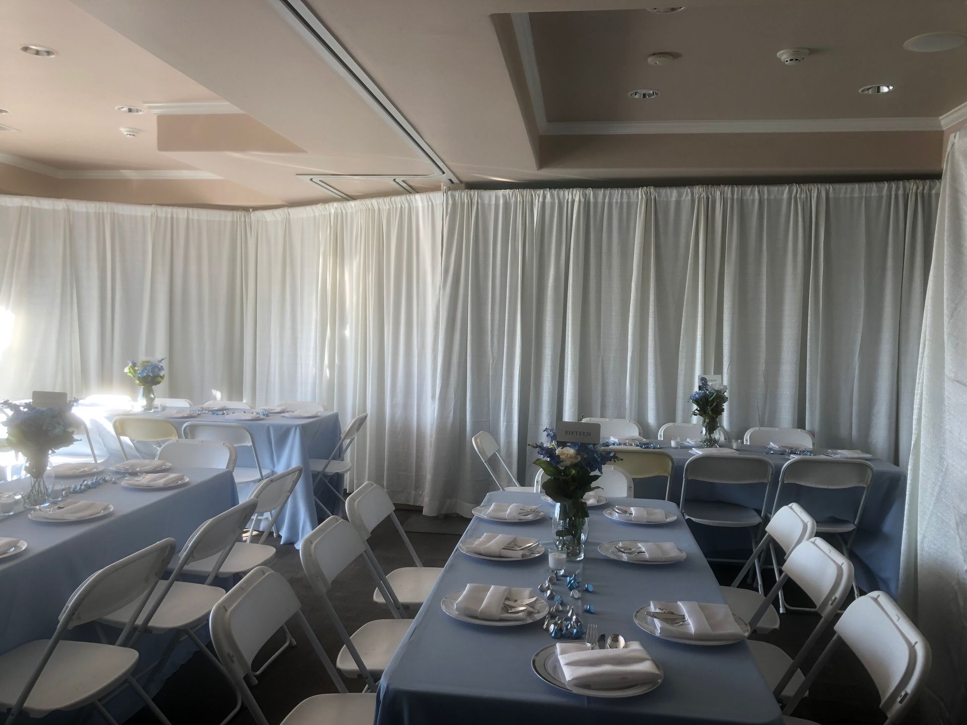 Tables set for a gathering in a room with white curtains. Blue tablecloths, white chairs, and floral centerpieces.