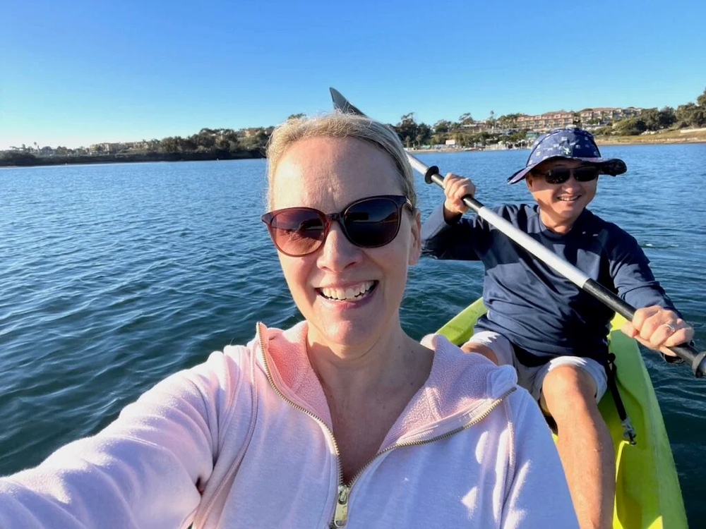 Woman in pink jacket, smiling, takes a selfie while kayaking with a person in a blue shirt on a lake.