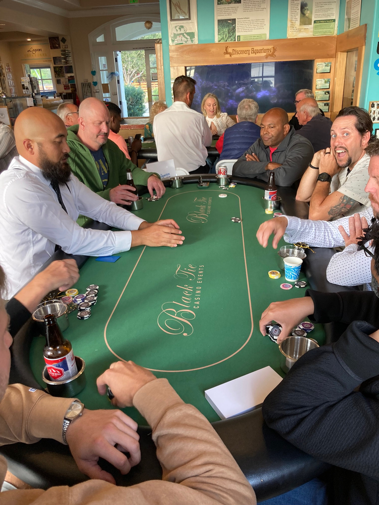 People playing poker at a table; some are holding cards, others smiling. Green felt, chips, and drinks are visible.