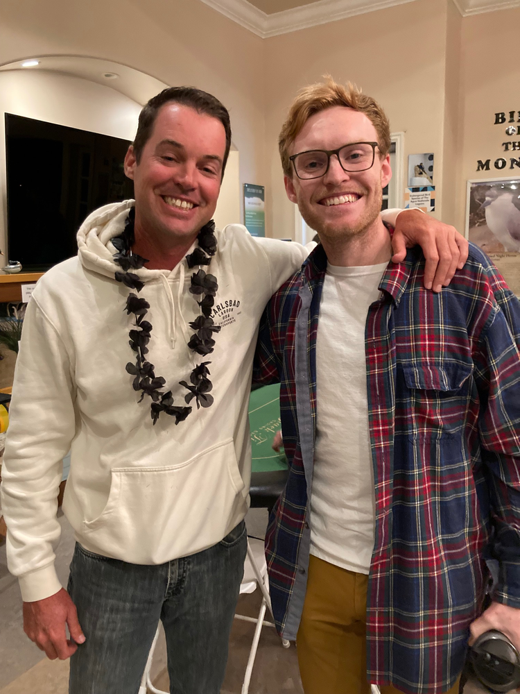 Two men smiling, one with a lei and white hoodie, the other with a plaid shirt, arm around the first man.
