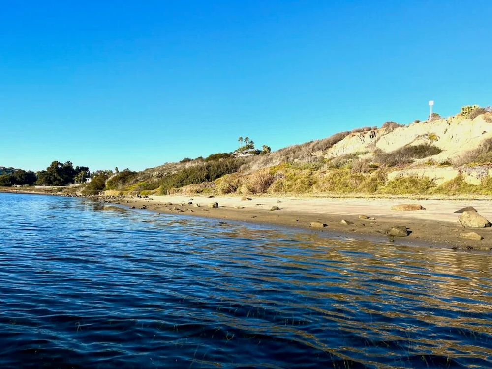 Coastal scene: blue water, sandy shore, tan bluffs under a clear blue sky.