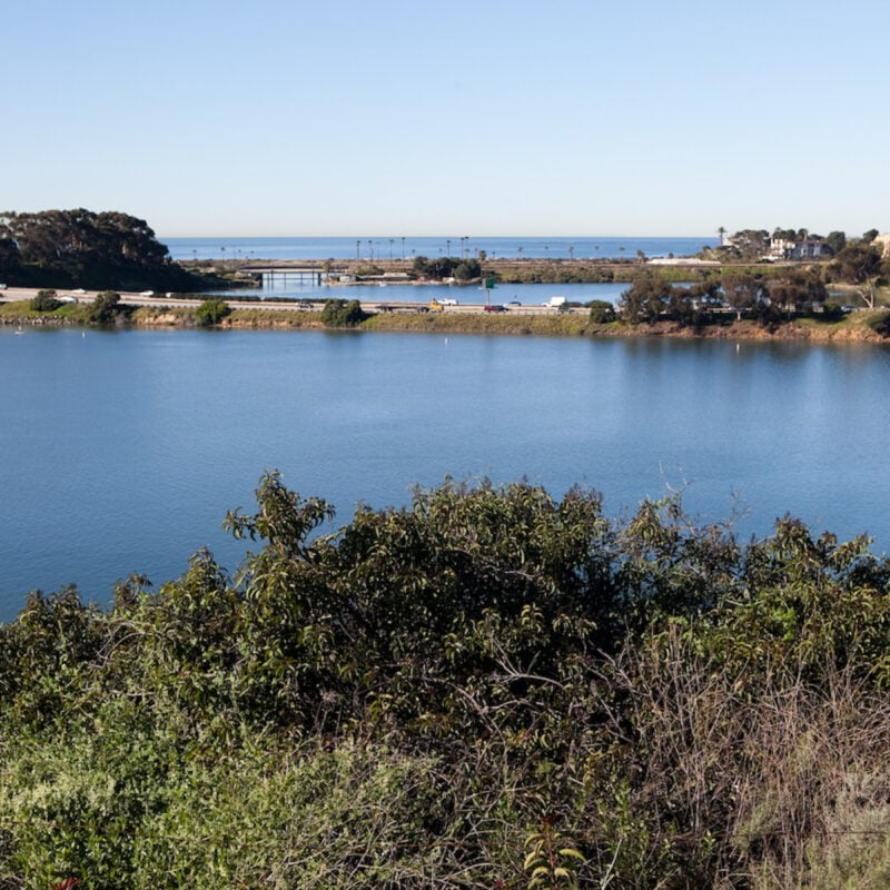 Blue lake with green bushes in the foreground, with structures and the ocean visible in the distance under a blue sky.