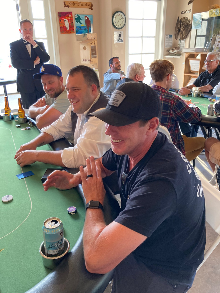 Men playing poker around a green table in a bright room. One man smiles, holding cards. Others watch.