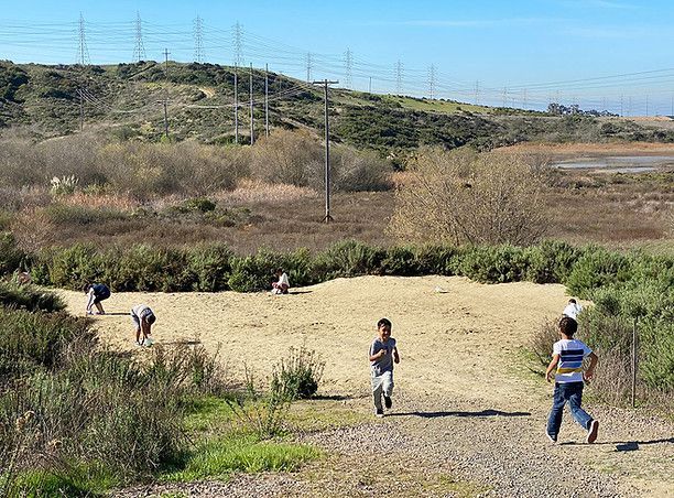 Children running on a dirt path in a sunny park with power lines and hills in the background.