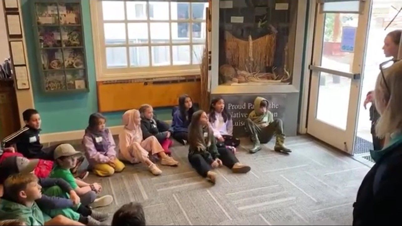 Children seated on floor listening to person near open doorway, possibly a museum setting.