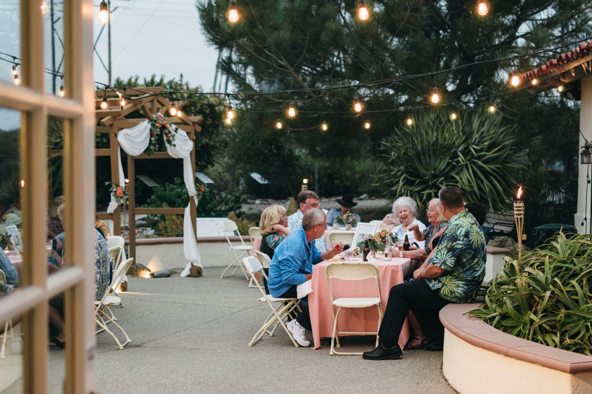 People at outdoor dinner party, pink tablecloth, string lights, patio setting.