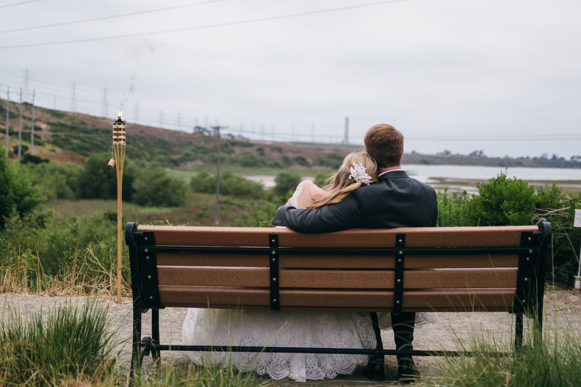 Couple in formal wear on a bench, backs to the camera, overlooking a natural landscape.