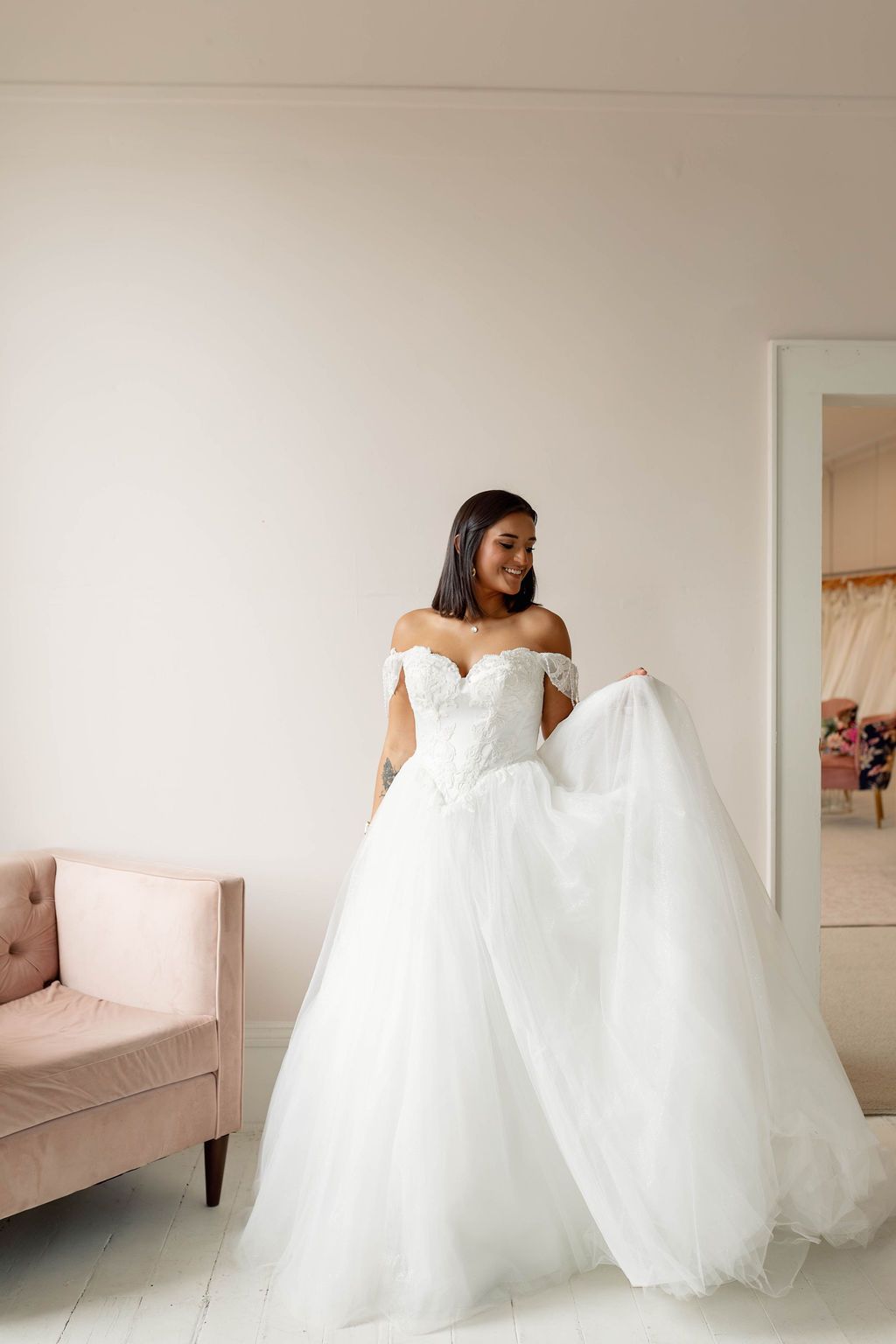 Woman in a white off-shoulder gown holding a long tulle train in a bright room