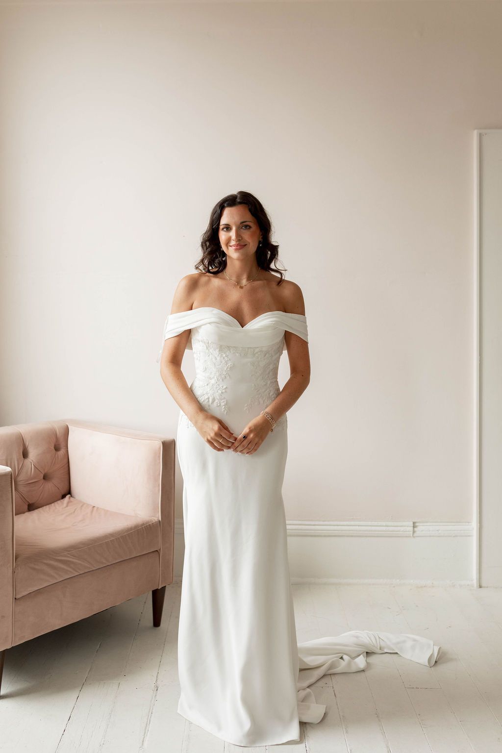 Woman in an off-shoulder white wedding dress standing in a bright room beside a blush pink sofa.