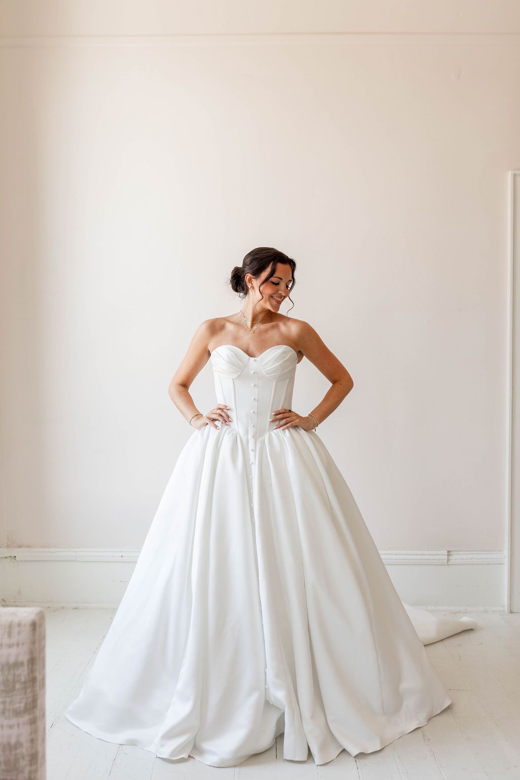 Bride in a strapless white gown standing in a bright room with hands on hips