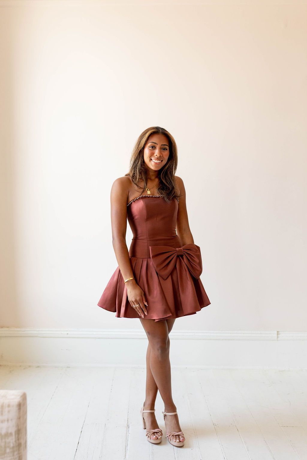 Woman in a rust-colored dress posing against a white wall and floor, wearing strappy heels.