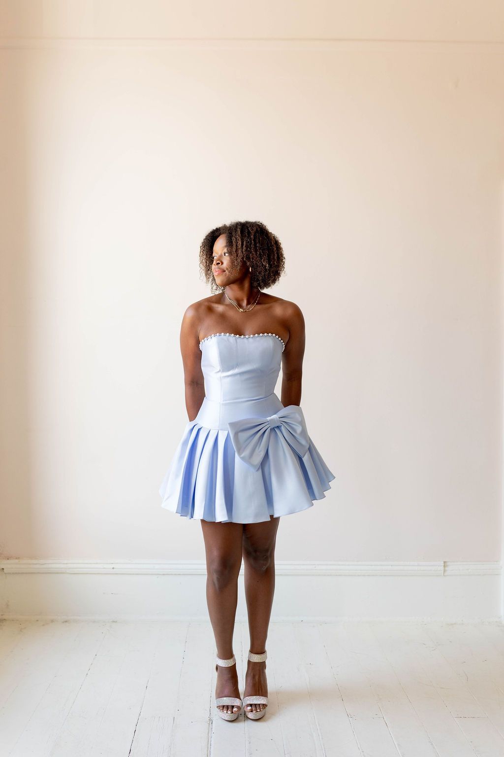 Woman in a light blue strapless dress and heels standing against a plain wall.