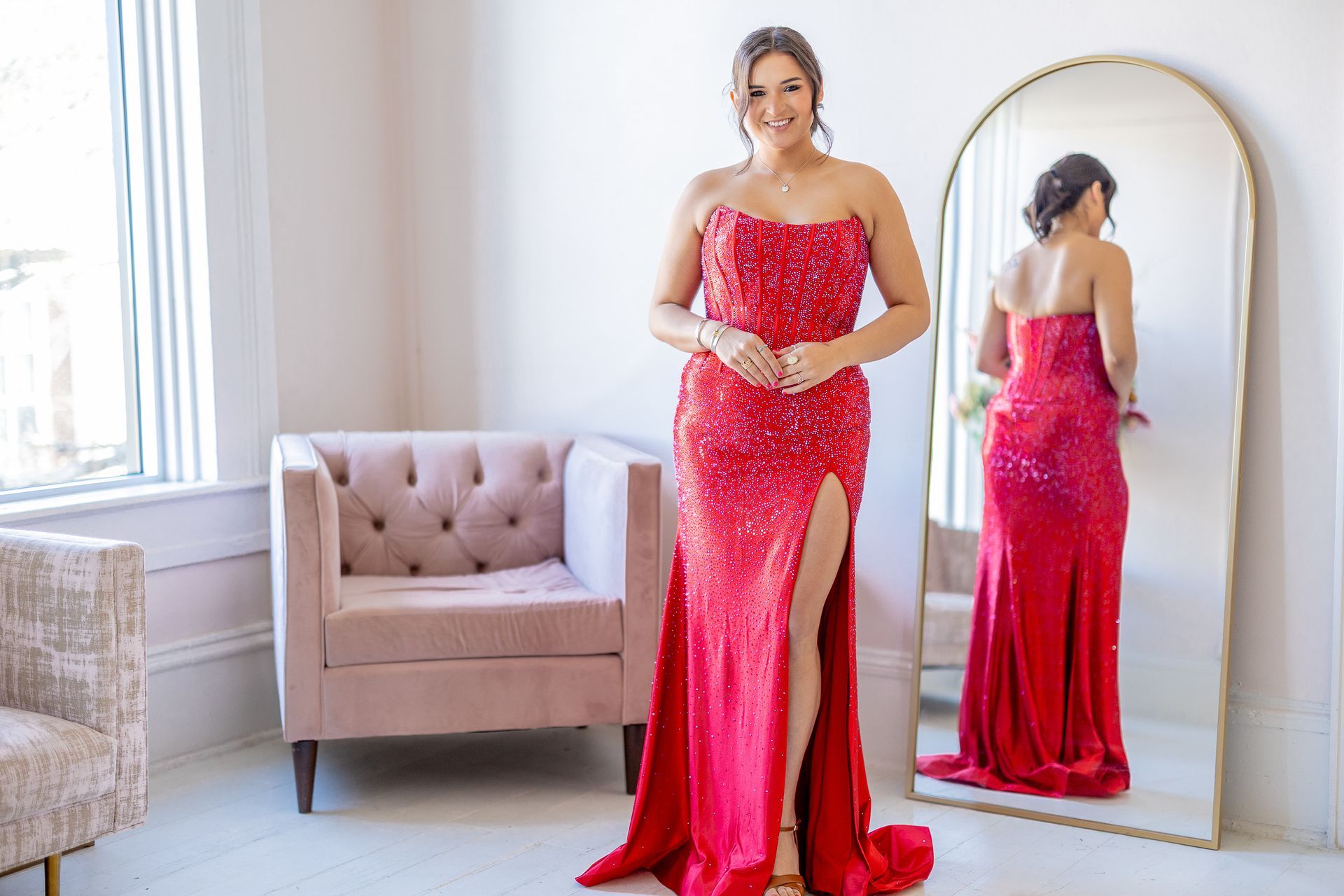 Woman in red sequined strapless gown, standing in front of a mirror and pink chair, by a window.