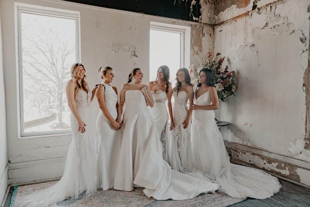 Six women in white gowns pose indoors with a distressed wall, natural light streaming through windows.