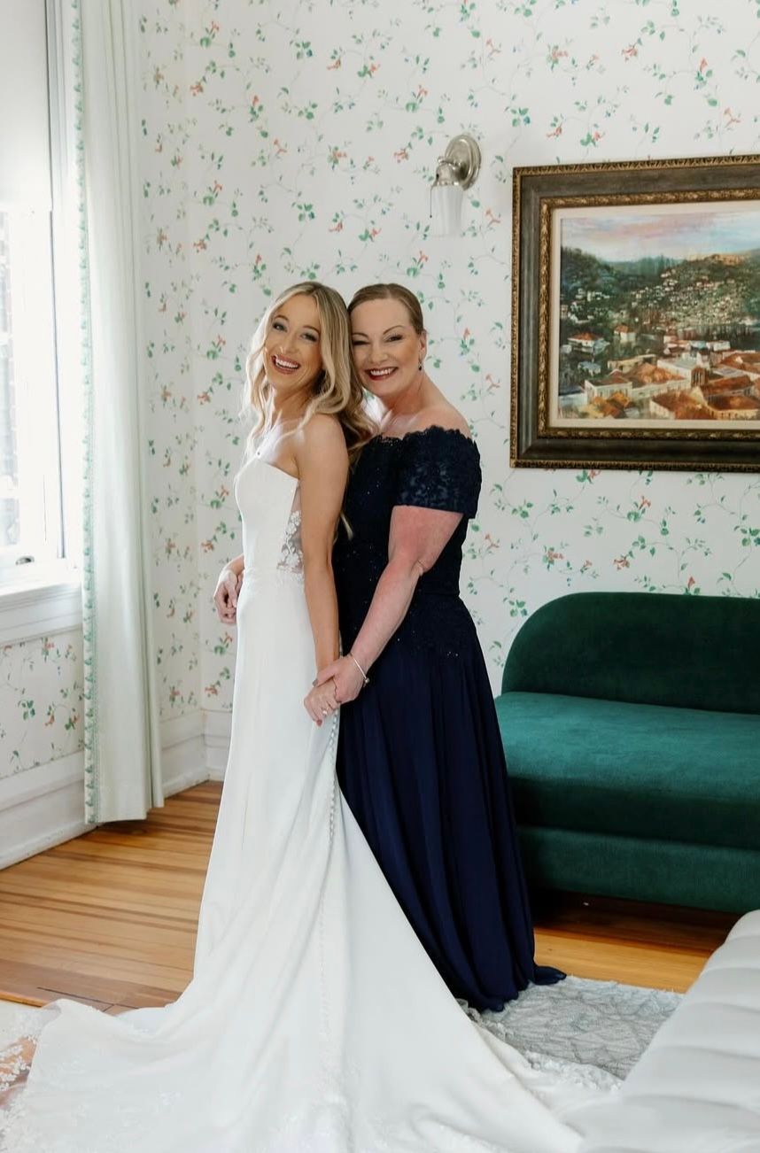 Bride and mother pose in a room. Bride in white gown, mother in navy dress, smiling.