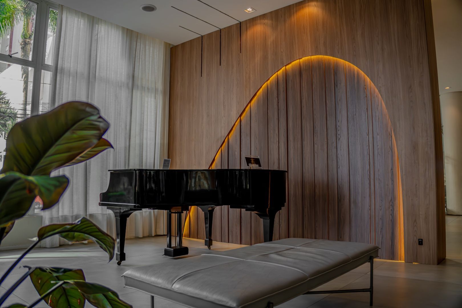 Piano de cauda preto em uma sala com uma parede de madeira curva, iluminada com luz quente, um banco e uma grande janela.