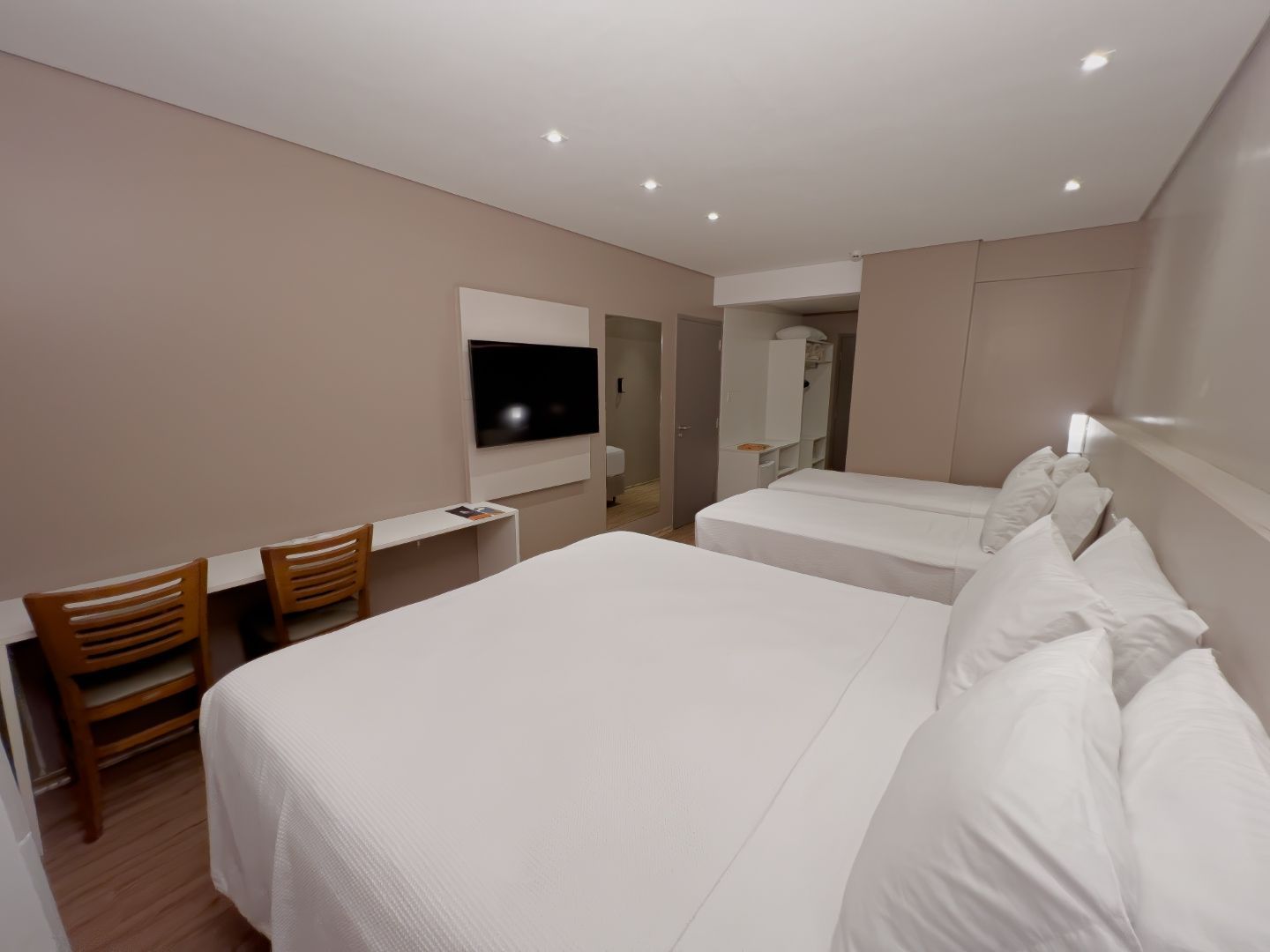 Quarto de hotel com duas camas, escrivaninha, cadeiras, TV e vista para o banheiro.