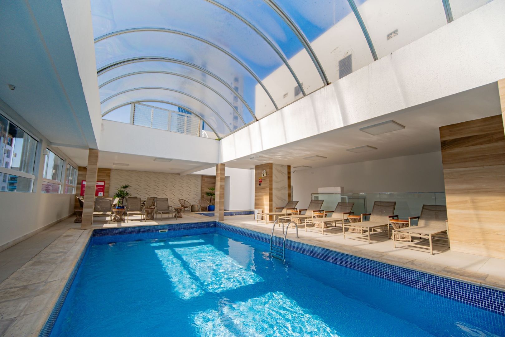 Piscina coberta com teto transparente, água azul e áreas de estar.