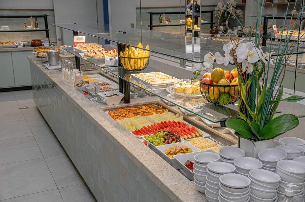 Buffet com comidas variadas, incluindo frutas, doces e pratos quentes, em um ambiente moderno e iluminado.