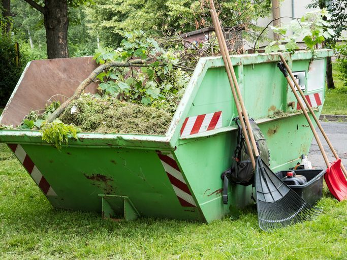 A green dumpster filled with leaves and branches is sitting in the grass.