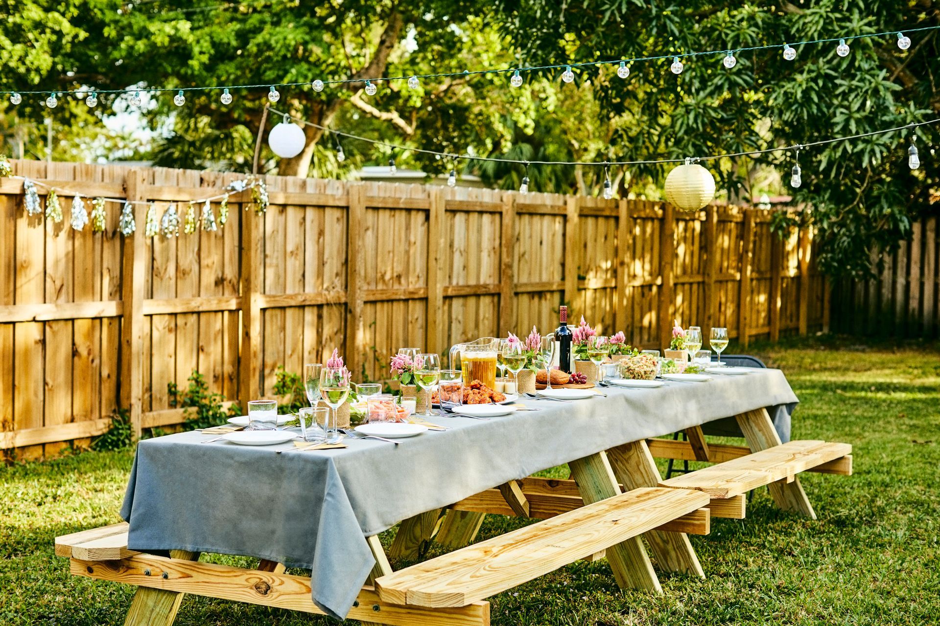 A long wooden picnic table is set for a party in a backyard.