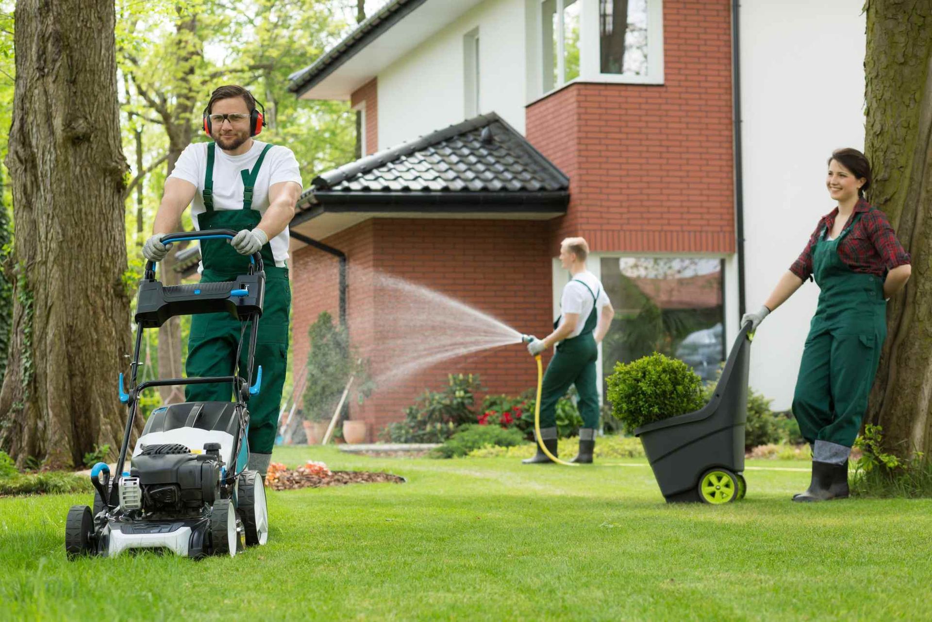 A man is mowing the grass in front of a house while a woman waters the lawn.